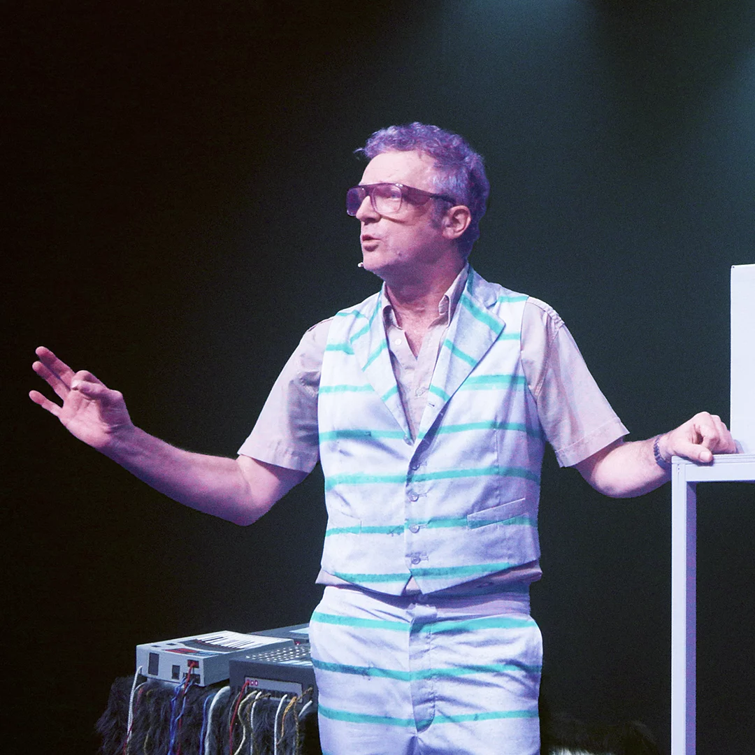 Speaker in a light-blue striped suit and oversized sunglasses delivering a presentation on stage, with audio equipment and cables visible nearby.