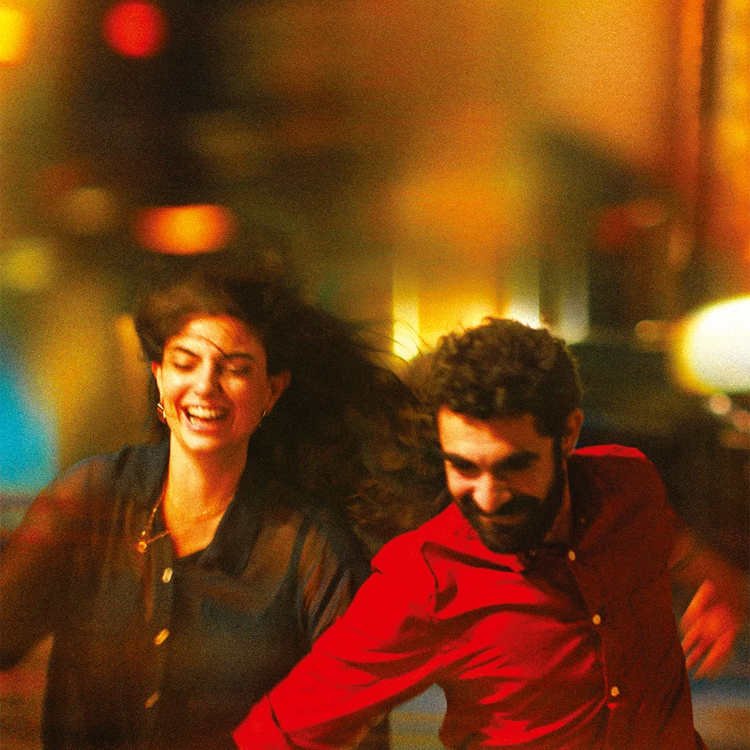 Two friends dancing joyfully in a vibrant city night scene, smiling as colorful lights blur around them.