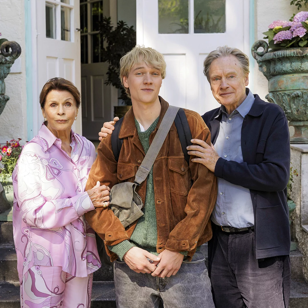 Three people stand on the front steps of a house: an elderly woman in a pink patterned blouse, a young man with a brown jacket and backpack, and an older man in a navy cardigan.