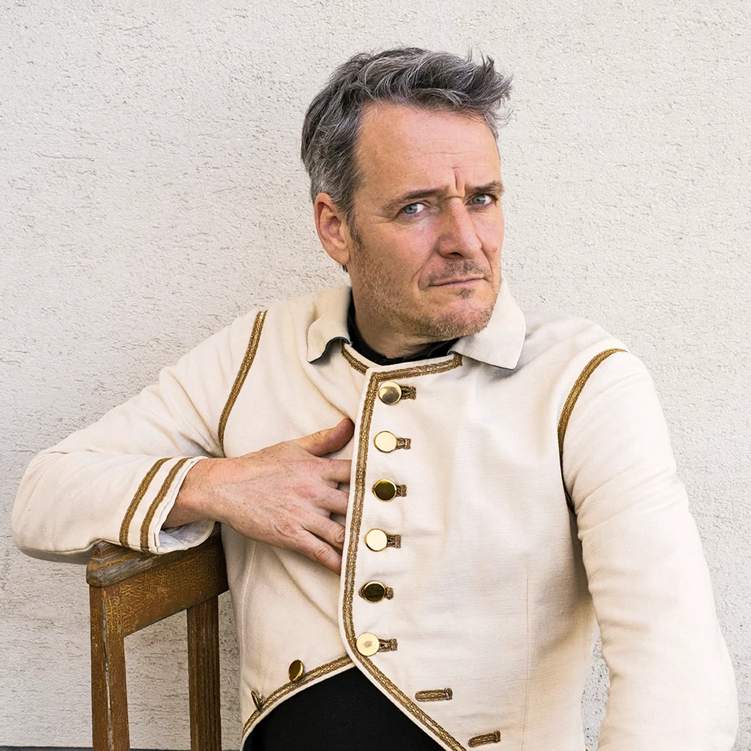 Portrait of a man in a cream-colored, military-style coat with gold buttons, seated on a wooden chair against a pale wall, looking to the side.