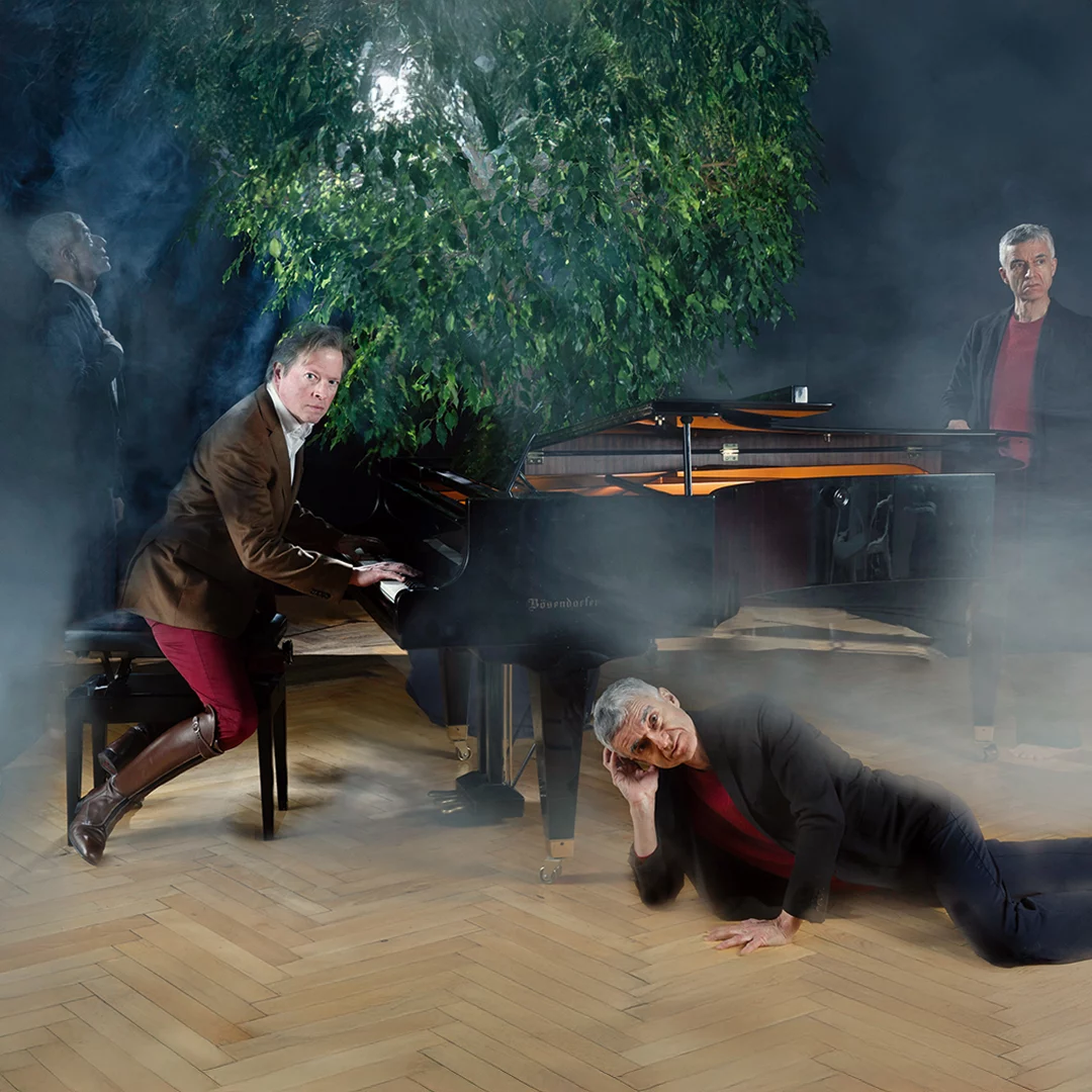 Four men around a grand piano in a smoky room; one plays, another lies on the floor, the others stand nearby.
