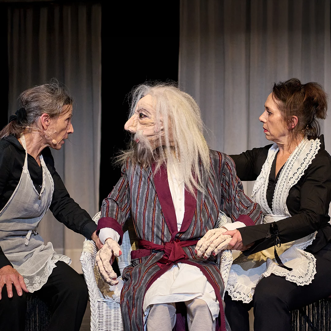 Theater scene with a bearded elderly man in a striped robe seated between two maids in black outfits and lace aprons.
