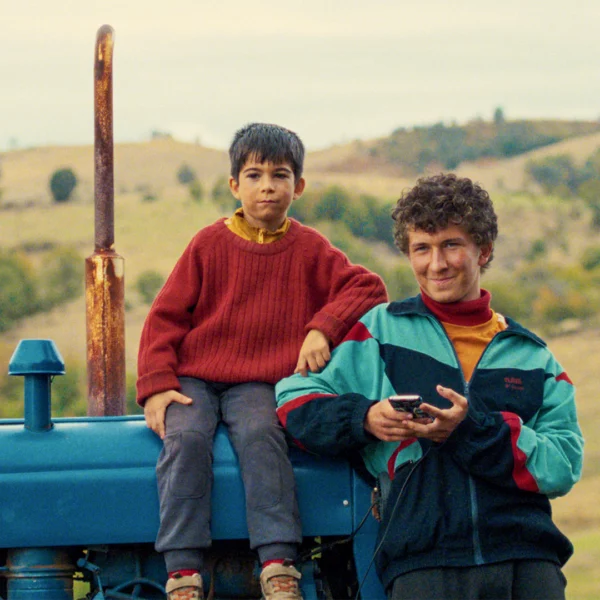Two boys sit on a blue tractor in a rural field; one in a red sweater, the other in a colorful jacket, holding a phone.