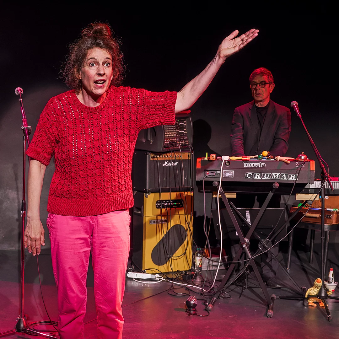 Female singer in a red knit top and pink pants raises her right arm on a stage, with a keyboardist and gear in the background.