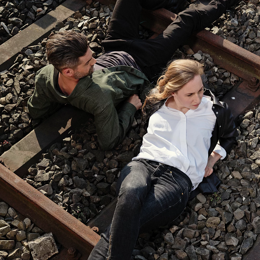 Two adults lying on railroad tracks among gravel, a man in a green shirt and a woman in a white blouse resting beside him.