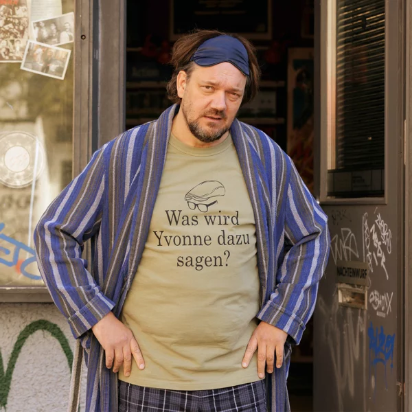 Man stands in a doorway wearing a blue striped robe and blue sleep mask, beige T-shirt reads: “Was wird Yvonne dazu sagen?” with a graffiti-covered storefront in the background.