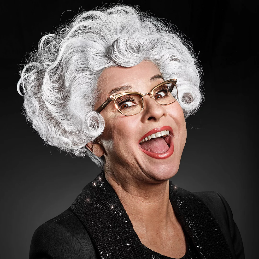 Joyful elderly woman with a voluminous white wig, glasses, and a glittery black outfit, mid-lake smile against a dark backdrop.