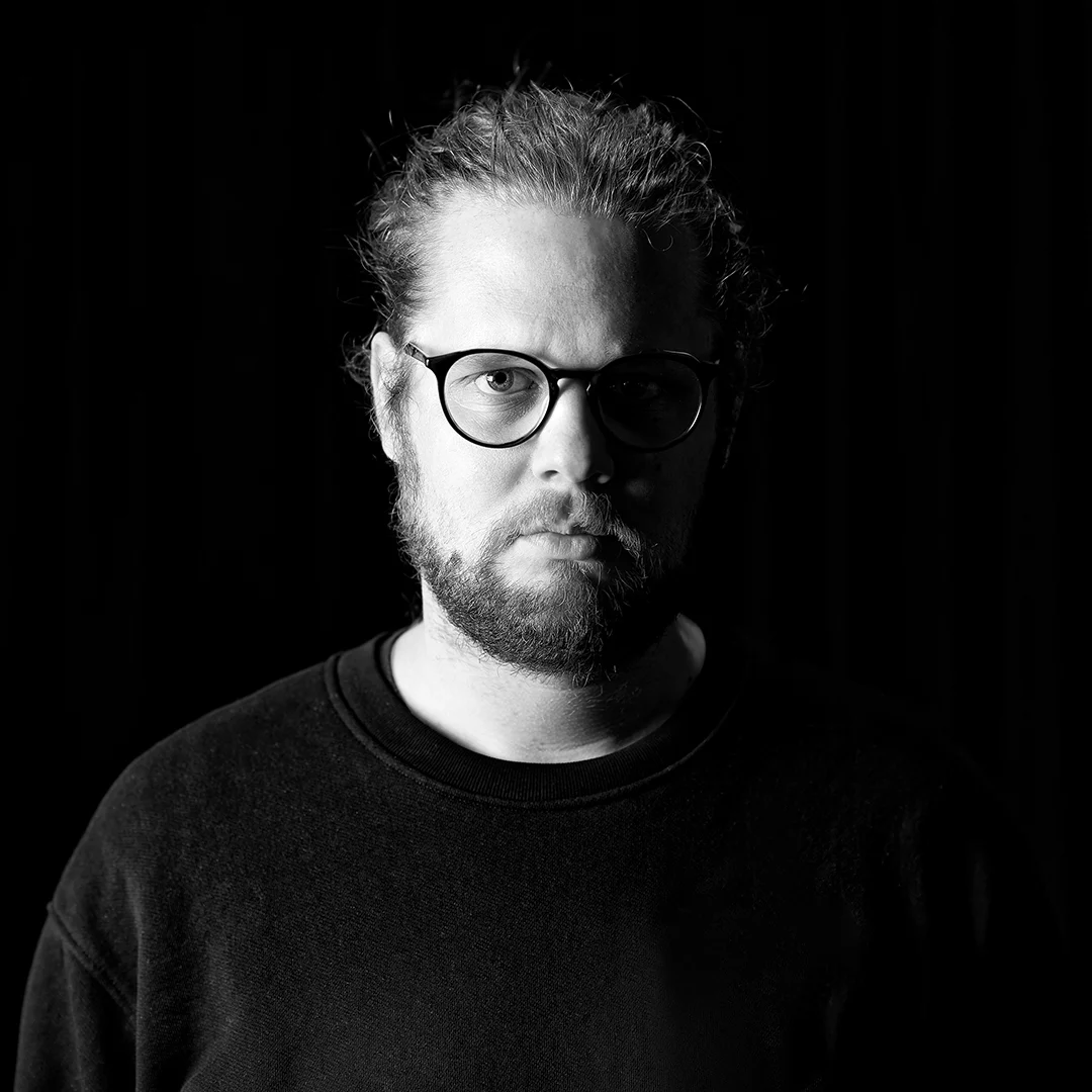 Black-and-white portrait of a man with round glasses and a beard, facing the camera against a dark background.
