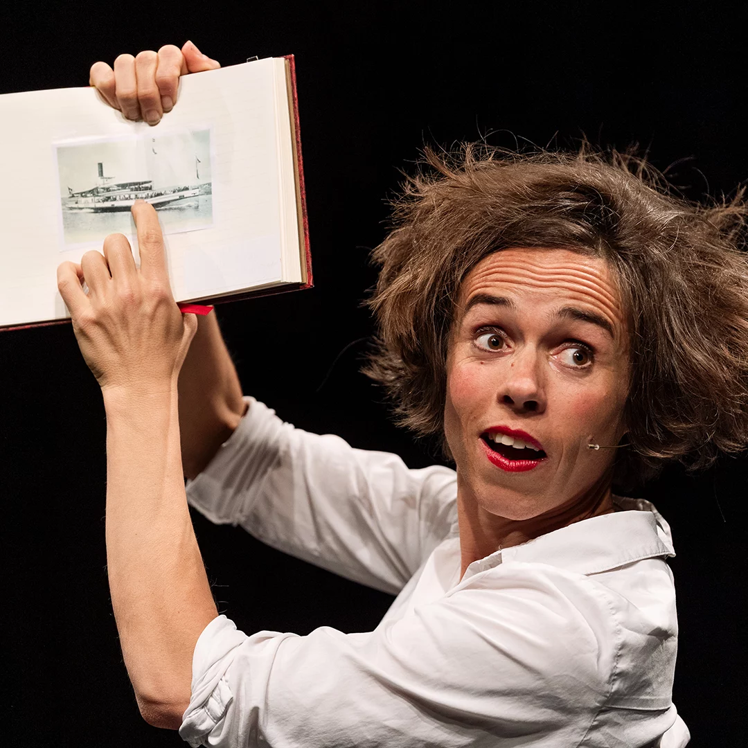Theatrical performer in a white shirt raises an open album, pointing to a photo with a surprised expression.