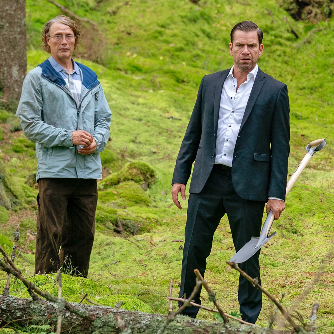 Two men stand in a mossy forest; one in a suit holding a shovel, the other in a light jacket, looking toward the camera.