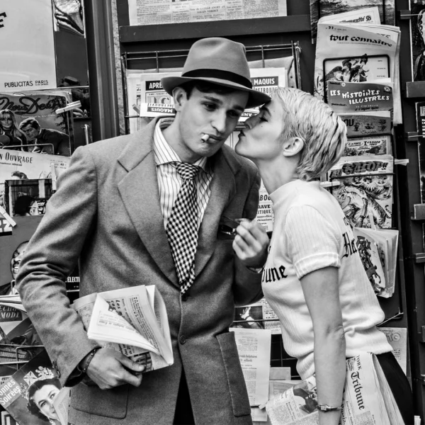 Man in a suit and fedora smokes a cigarette as a woman with short hair leans in to kiss him in a busy newspaper stand filled with magazines and papers.