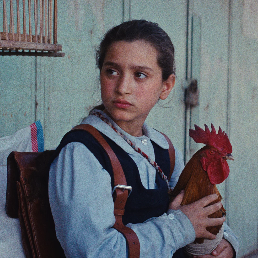 A young girl with a backpack gently holding a rooster as she sits against a pale wall, looking to her left.