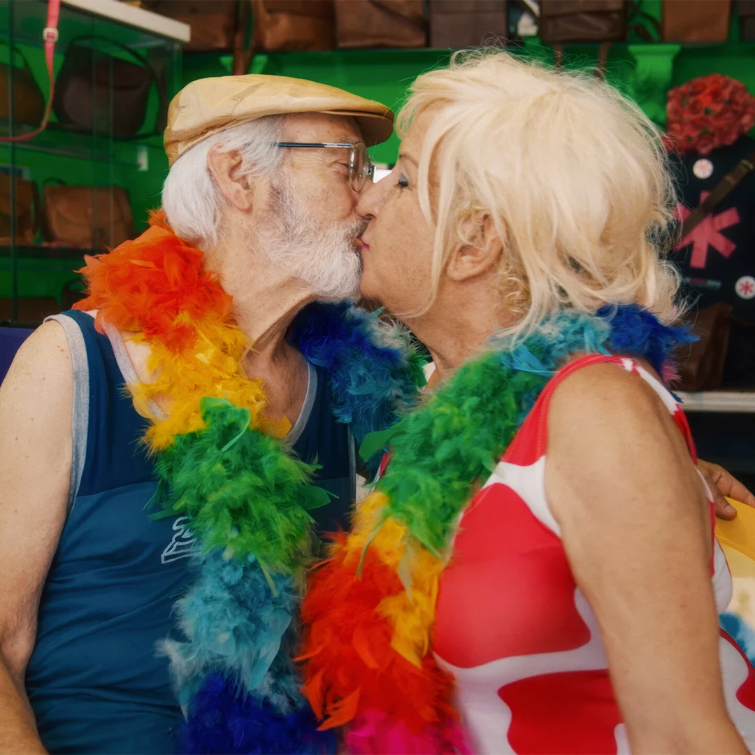 Two older adults kiss at a festive event, wearing rainbow feather leis and bright clothing.