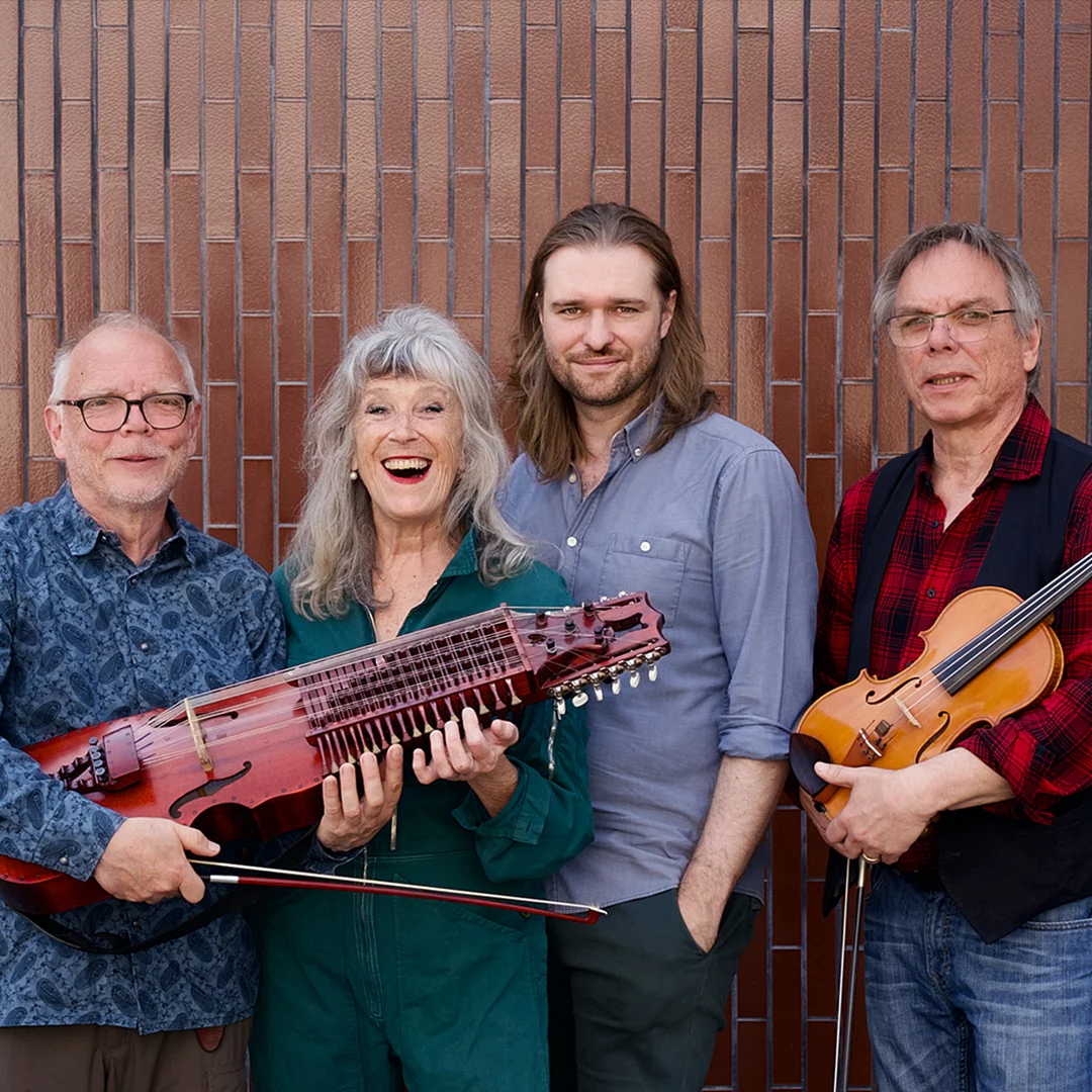 Four musicians posing against a brick wall; the woman in green center holds a long red stringed instrument while two men hold a bow and a violin, all smiling.