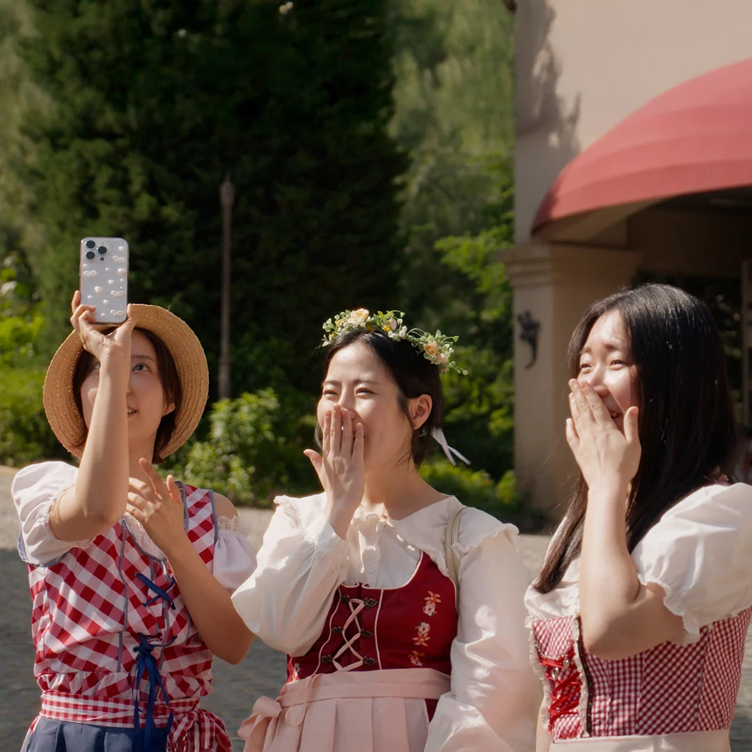 Three friends in festive dirndl-style dresses laugh and pose for a selfie outdoors, one wearing a flower crown.