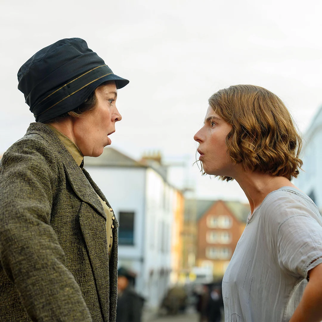 Two women face off and argue on a city street, with buildings in the background.