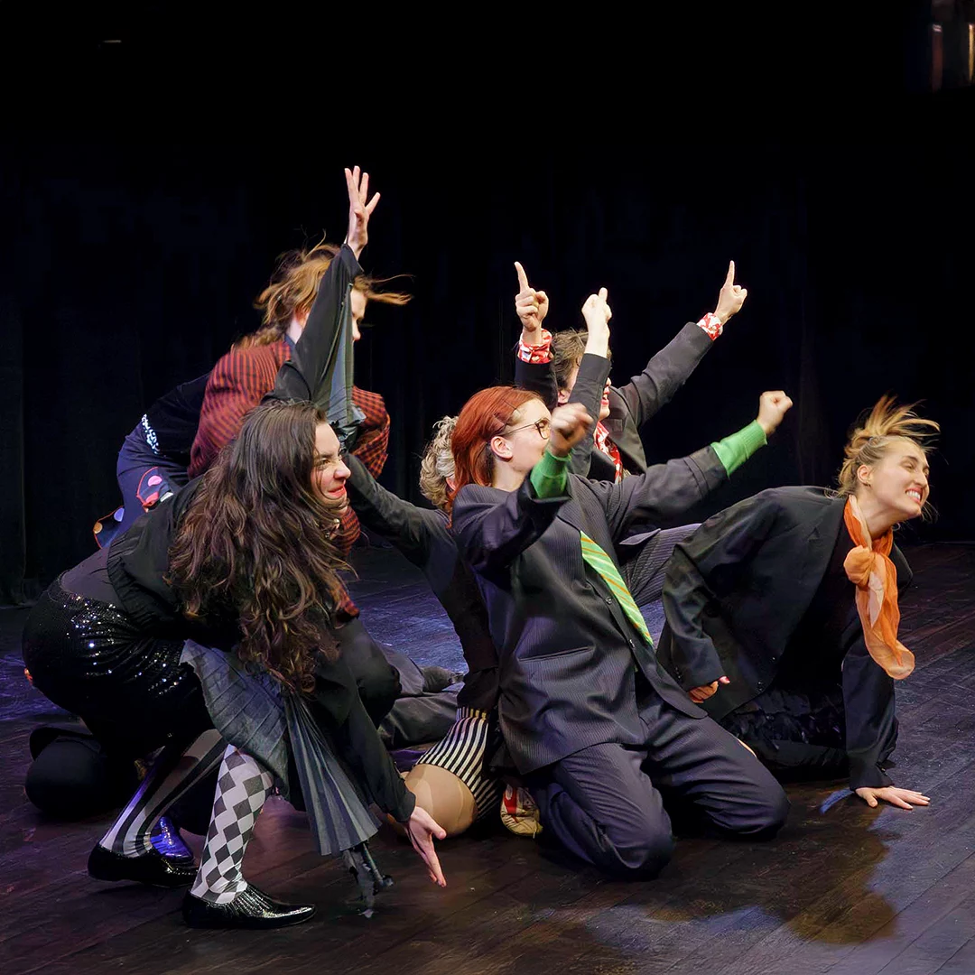 Group of performers on a dark stage striking a dynamic pose with raised arms and varied outfits.