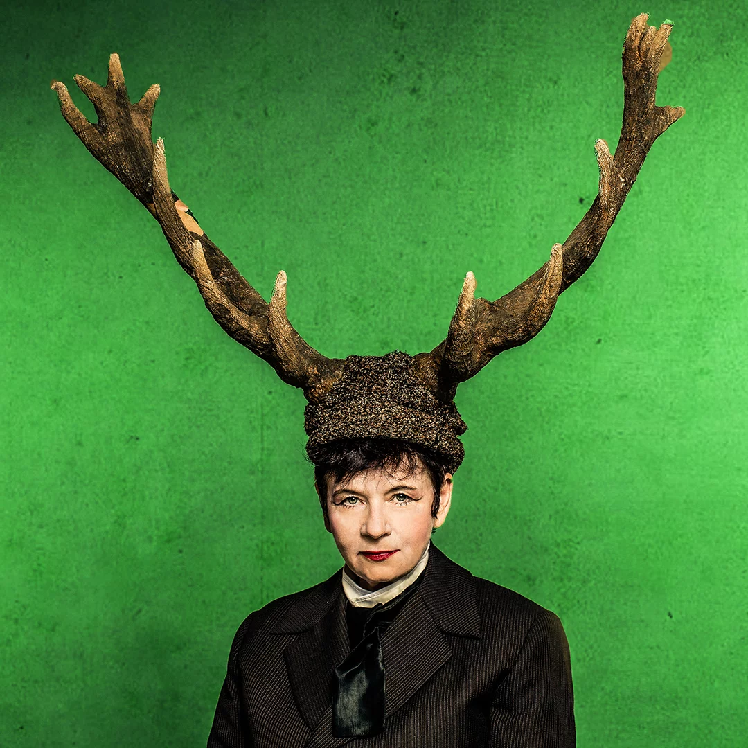 Portrait of a woman in a dark suit wearing a large deer antler headpiece against a bright green background.